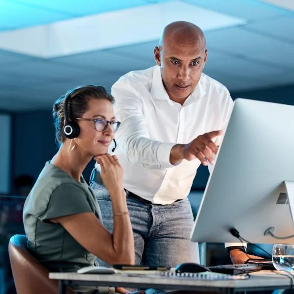 A man and woman collaborate on a computer in a call center office, focusing on workforce management tasks.