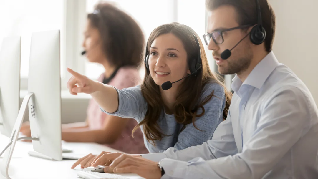 A man and woman in headsets collaborate on computers in a call center environment, focusing on workforce management tasks.