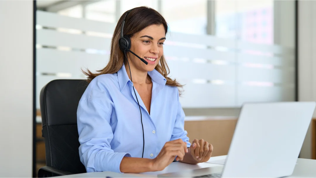 A woman in a headset sits at a desk with a laptop, engaged in remote customer service.